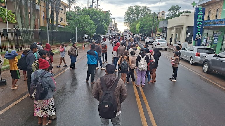 Grupo de indígenas descalzos y con mochilas, escuchando a mujer que habla en una calle con policías y vehículos al fondo.