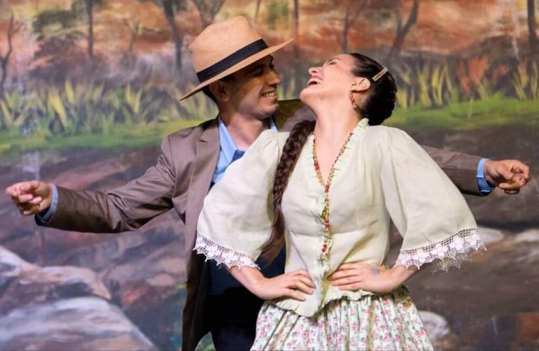 Hombre en traje gris y sombrero claro junto a mujer en vestido tradicional sonríen y bailan en un fondo natural.