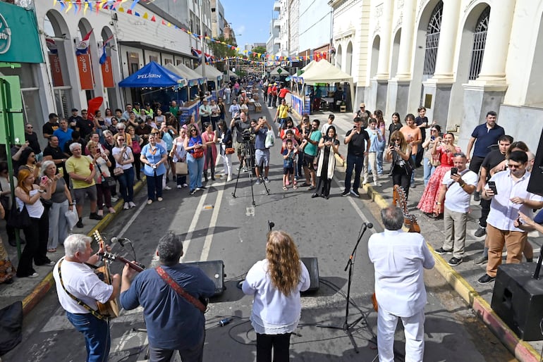 Cinco músicos tocan en una celebración al aire libre, rodeados de una multitud de personas con decoraciones festivas.