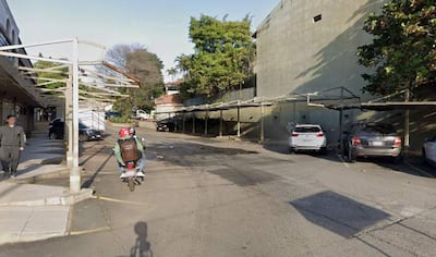Hombre con ropa oscura camina mientras un motociclista con casco rojo pasa en una calle urbana con árboles de fondo.