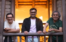 Tres hombres sonrientes en balcón de madera; uno con camiseta morada y gafas grandes, gesto relajado y amigable.