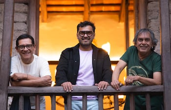 Tres hombres sonrientes en balcón de madera; uno con camiseta morada y gafas grandes, gesto relajado y amigable.