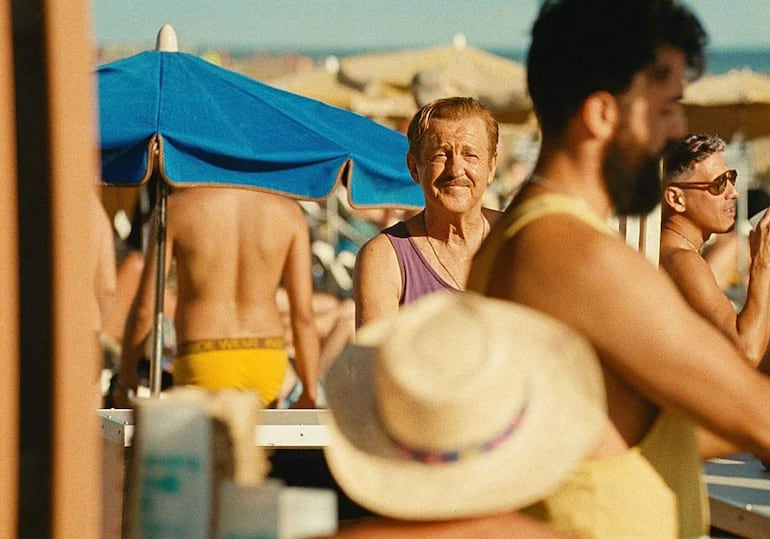 Hombre de edad avanzada con bigote y camiseta morada sonríe en la playa, rodeado de bañistas y sombrillas.