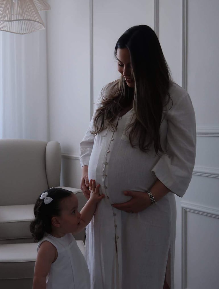 Mujer embarazada de cabello lacio en blusa clara mira con ternura a su hija con cabello rizado y moño blanco, en ambiente luminoso.