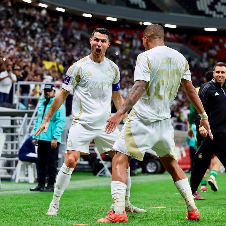 Cristiano Ronaldo, con camiseta blanca y detalles dorados, celebra con otro jugador, levantando los brazos en un estadio lleno de aficionados.