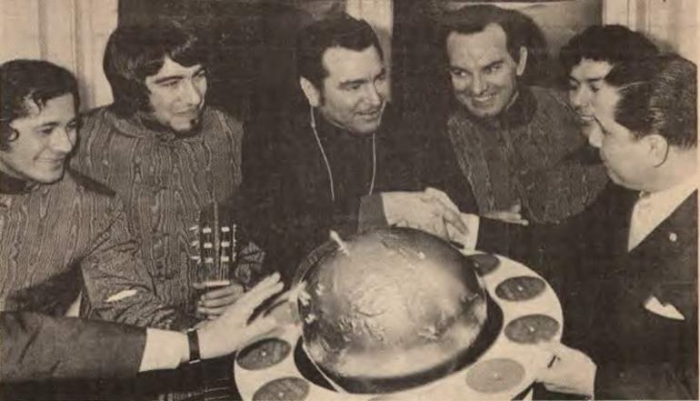 Fausto Franco y Adrián Barreto se dan la mano en un interior, rodeados de otros hombres, uno sostiene una guitarra.