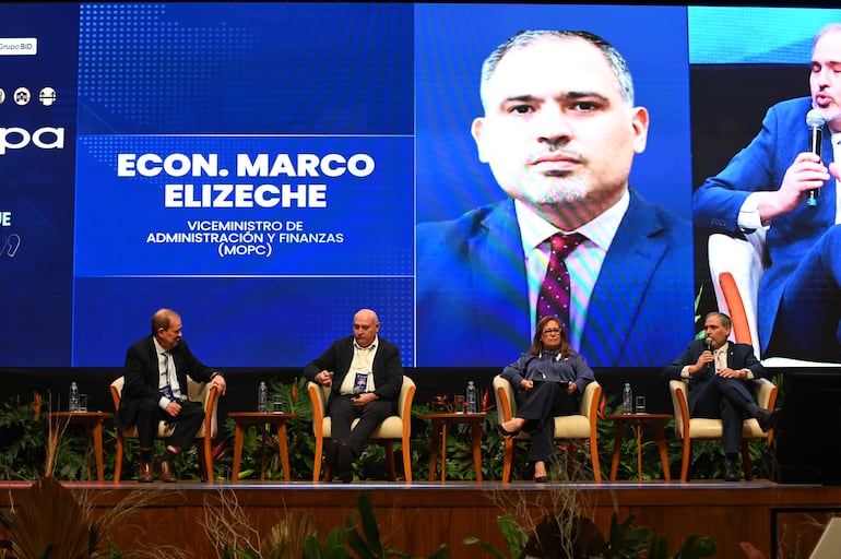 Marco Elizeche en el podio, vestido con traje oscuro, César Barreto a su lado, ambos escuchando atentamente en un evento con elegante decoración.