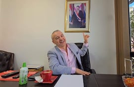 Daniel Centurion en blazer gris y camisa rosa, sonríe mientras señala un retrato de Mario Abdo Benítez en la pared.