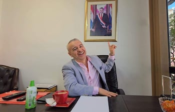 Daniel Centurion en blazer gris y camisa rosa, sonríe mientras señala un retrato de Mario Abdo Benítez en la pared.