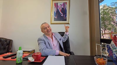 Daniel Centurion en blazer gris y camisa rosa, sonríe mientras señala un retrato de Mario Abdo Benítez en la pared.