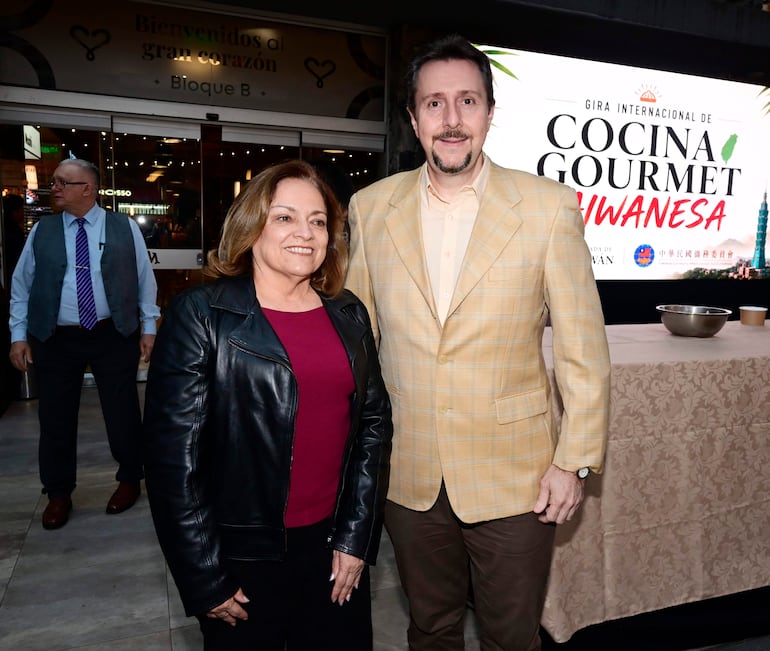 Mujer en camiseta burdeos y chaqueta negra junto a hombre en chaqueta de cuadros amarillos, ambos sonrientes en evento culinario.