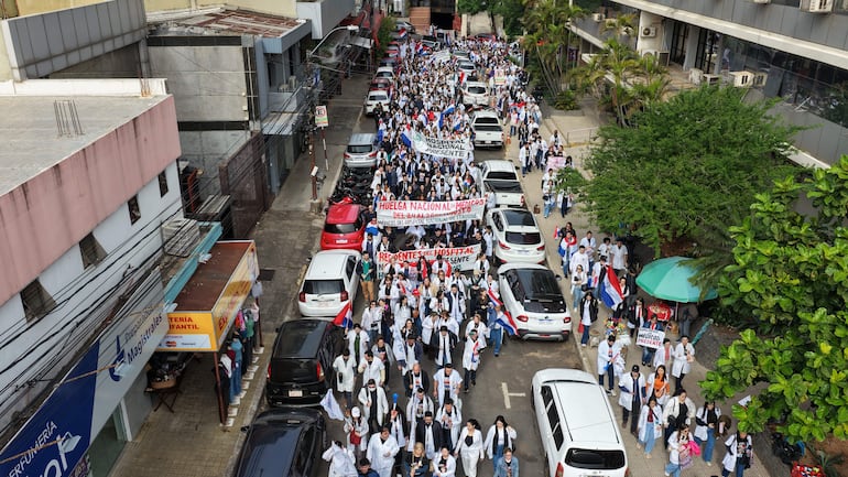 Cientos de profesionales de la salud en batas blancas marchan enérgicamente, sosteniendo pancartas, entre banderas de Paraguay en un entorno urbano.