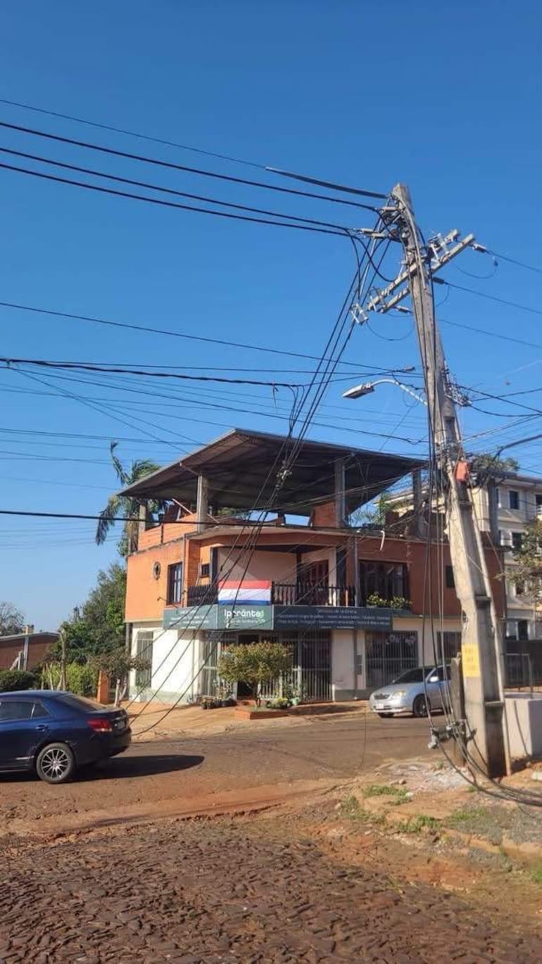 Edificio de ladrillo con balcón y bandera paraguaya, postes y cables eléctricos visibles en un barrio tranquilo.