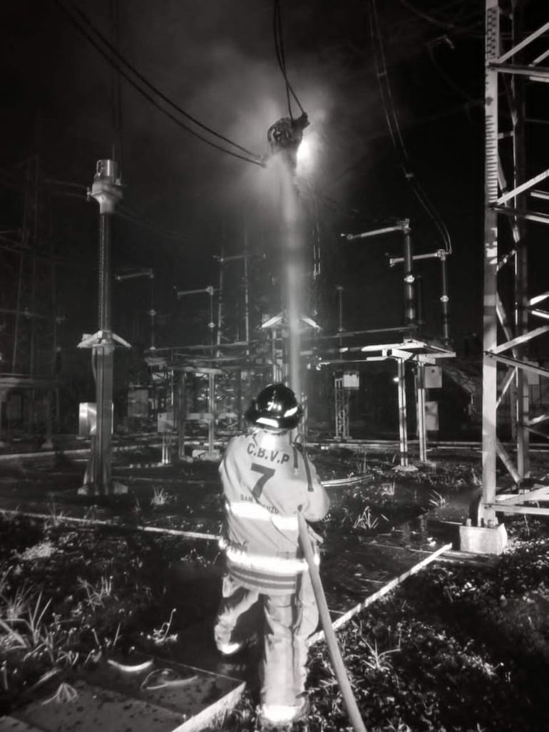 Bombero con chaqueta de C.B.V.P. apunta una manguera hacia un incendio en un entorno industrial oscuro y lleno de humo.