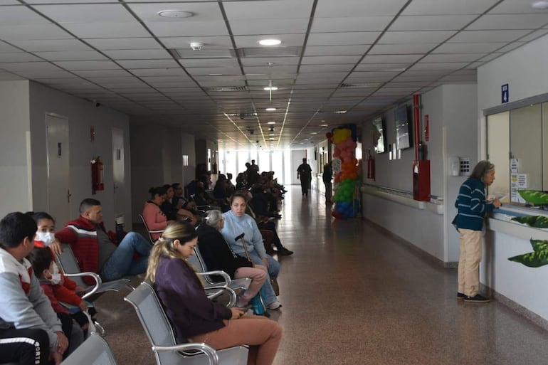 Mujer en mostrador interactuando con personal, mientras otros esperan sentados en sillas metálicas.