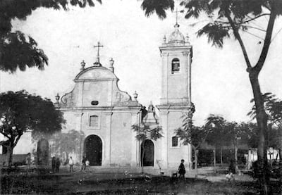 Iglesia colonial blanca con campanario y cruz, rodeada de árboles en un día despejado.