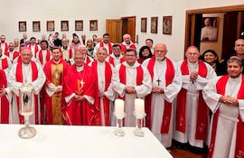 Grupo de sacerdotes y obispos en vestimenta litúrgica roja y blanca, con mesa central y figura religiosa decorando el fondo.