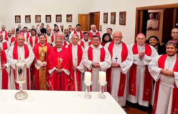 Grupo de sacerdotes y obispos en vestimenta litúrgica roja y blanca, con mesa central y figura religiosa decorando el fondo.