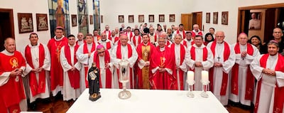 Grupo de sacerdotes y obispos en vestimenta litúrgica roja y blanca, con mesa central y figura religiosa decorando el fondo.