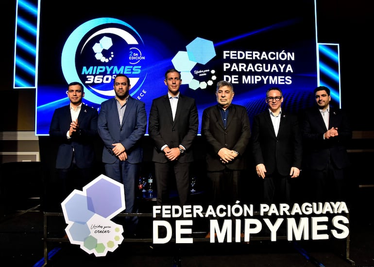 Seis hombres en trajes formales participan en un evento, con el logo de MIPYMES de fondo y copas sobre la mesa.