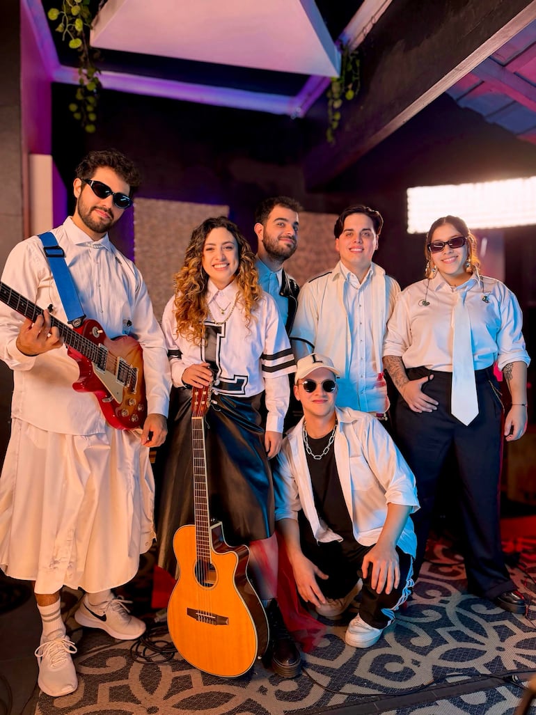 Luce en el centro sosteniendo una guitarra acústica, rodeada de músicos con diferentes estilos, en un ambiente decorado y alegre.