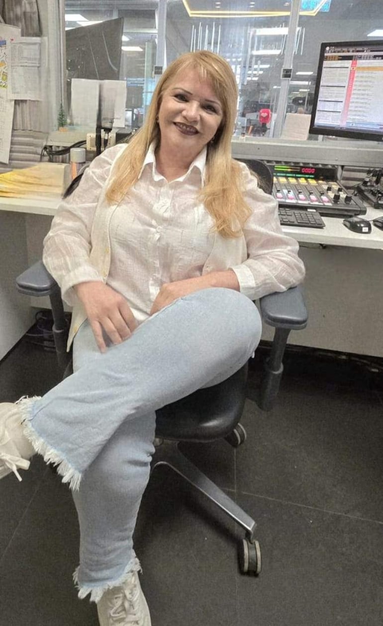 Mujer de cabello rubio y largo, sonriente, vestida con camisa blanca y chaqueta, sentada en oficina moderna.