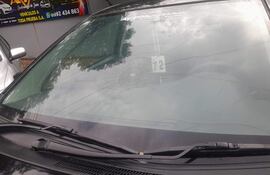Automóvil con parabrisas dañado y grietas visibles, en un entorno urbano con otros vehículos y un negocio al fondo.