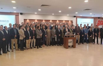 Grupo de empresarios en ambiente formal, escuchando a un orador en traje mientras se ven atentos y serios.