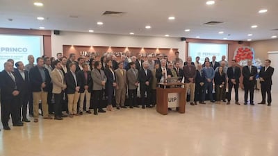 Grupo de empresarios en ambiente formal, escuchando a un orador en traje mientras se ven atentos y serios.