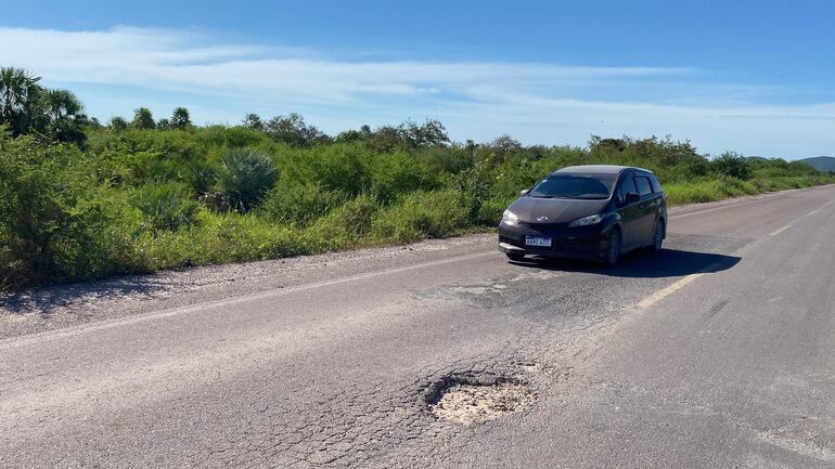 Automóvil oscuro en carretera rural rodeado de vegetación, con un bache visible en la superficie del asfalto.