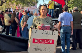 Mujer con camiseta verde y cabello castaño sostiene cartel que dice 'LA SALUD ES UN DERECHO NO UN PRIVILEGIO', rodeada de manifestantes.