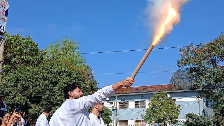 Grupo de jóvenes con camisas blancas, uno sostiene un tubo con fuego, en un evento al aire libre con árboles y edificios de fondo.