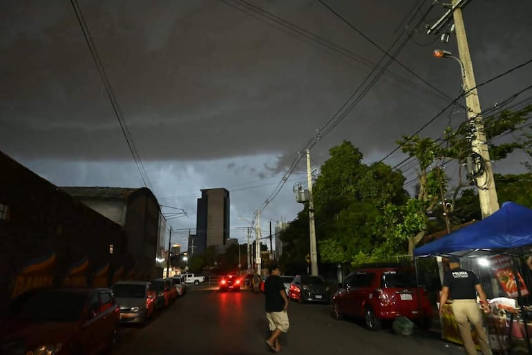 Hombre en camiseta de policía observa desde un puesto azul en una calle oscura y lluviosa, con tormenta inminente.