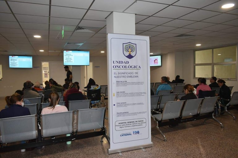Ambiente hospitalario moderno con pacientes en espera; hombre de pie frente a un banner informativo sobre oncología.