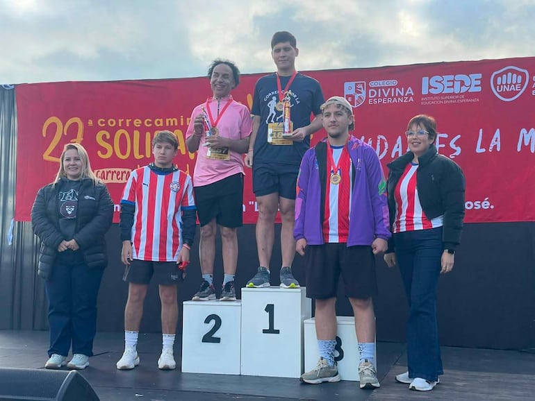 Hombre en camiseta negra sostiene trofeo en podio, mientras otros corredores celebran en niveles inferiores con sonrisas y trofeos.