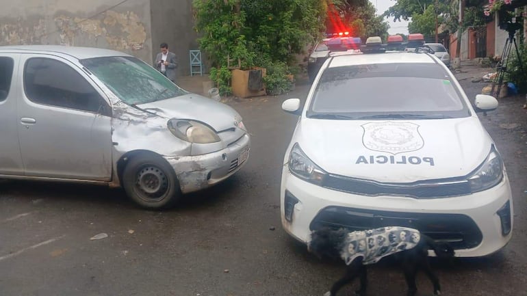 Vehículo policial blanco patrullando junto a coche plateado dañado. Persona formal revisa su celular en una calle con vegetación.