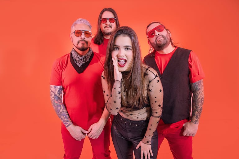 Cuatro integrantes de Cielo Ámbar posan juntos, dos hombres de camiseta roja, mujer en atuendo negro y chaleco negro en fondo naranja.