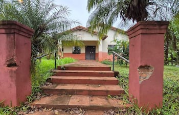 Estructura de salud blanca y naranja en mal estado, rodeada de vegetación, con escaleras desgastadas y sin personas visibles.