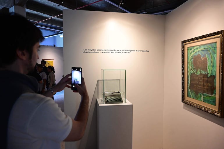 Hombre con camiseta blanca y chaleco oscuro sostiene un teléfono móvil, capturando imágenes de una máquina de escribir en exhibición.