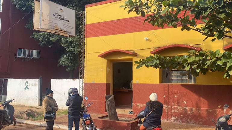 Mujer con abrigo beige y sombrero conversa con hombre de espaldas, mientras otra mujer observa, en un entorno soleado junto al Correo Paraguayo.