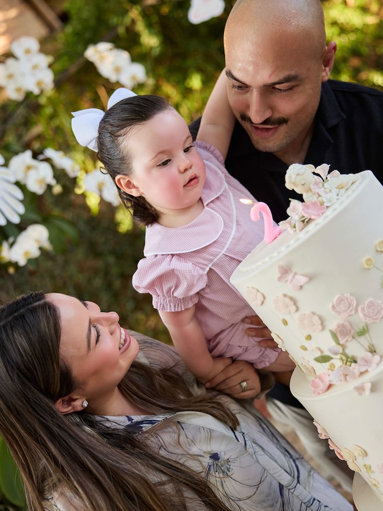 Hombre calvo y con bigote sostiene a una niña en vestido rosa, sonriente, durante su cumpleaños, rodeados de flores y pastel.