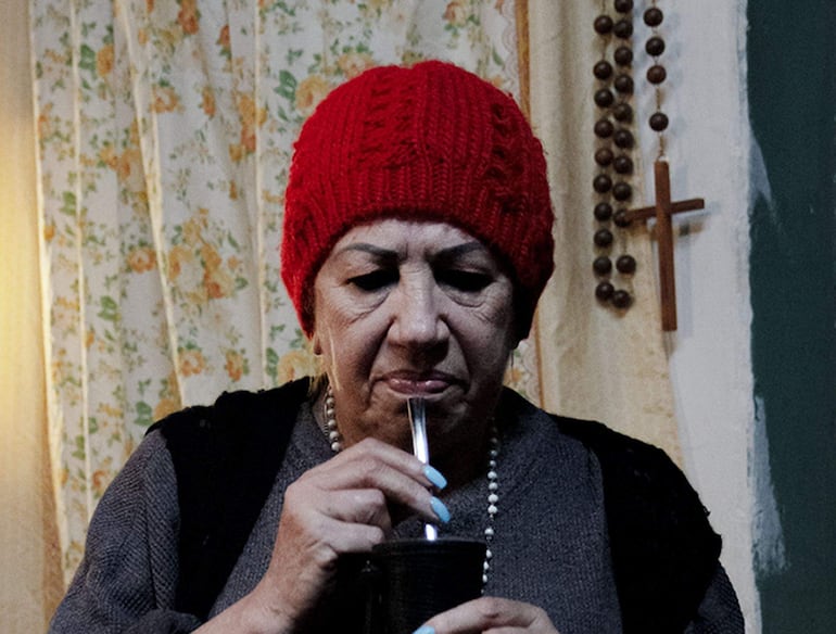 Mujer con gorro rojo bebe mate, vistiendo camiseta gris y chaleco negro, en un entorno decorado con flores y elementos religiosos.