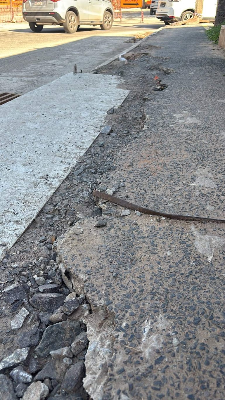 Calle en mal estado con concreto roto y escombros, sin personas visibles y vehículos estacionados al fondo.