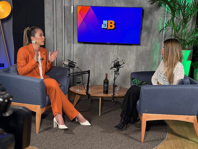 Jessica Torres en traje naranja gesticulando y escuchando, junto a Belén Bogado  con blusa blanca en un set con micrófonos y decoración moderna.