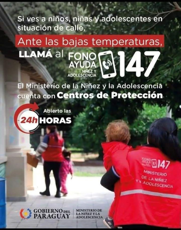 Dos trabajadores en uniformes rojos del Ministerio de la Niñez sostienen y ayudan a un niño en un parque durante la noche.