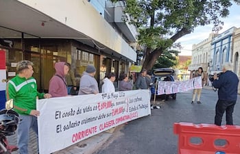 Grupo de seis manifestantes sosteniendo pancartas sobre salario mínimo; hombre con saquito verde al frente. Edificio histórico detrás.