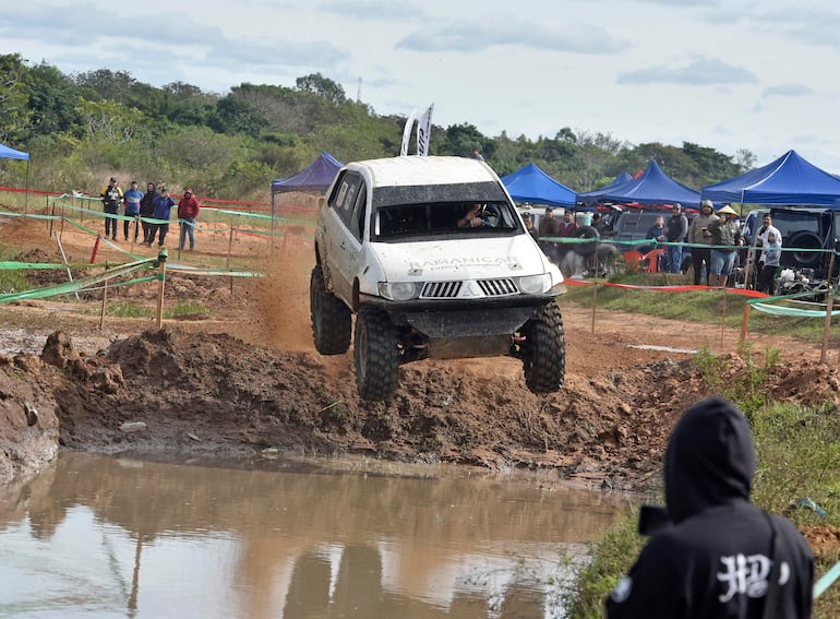 Vehículo SUV blanco saltando sobre el barro, rodeado de espectadores con ropa casual y carpas azules al fondo.