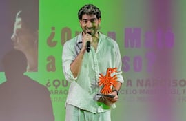 Diro Romero sonriendo, vistiendo camisa de rayas y pantalones claros, con un trofeo naranja en un escenario decorado.