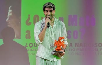 Diro Romero sonriendo, vistiendo camisa de rayas y pantalones claros, con un trofeo naranja en un escenario decorado.