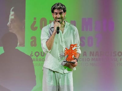 Diro Romero sonriendo, vistiendo camisa de rayas y pantalones claros, con un trofeo naranja en un escenario decorado.
